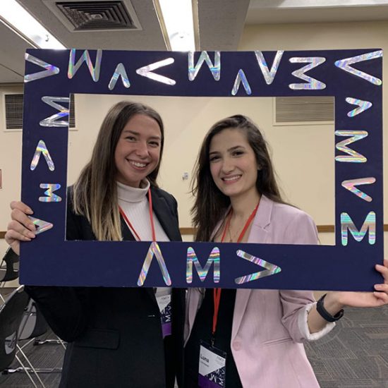 Two female students in business attire smiling, together holding a frame around their faces with the 