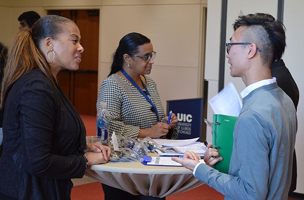 A black woman stands at a tall table talking to a younger man while a woman in the background stands behind the table talking to someone else.