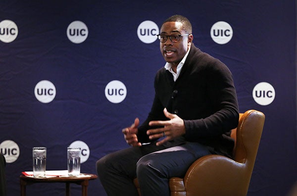 A Black male sitting in a chair, speaking and gesturing with his hands.