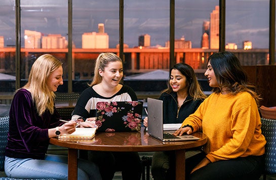 Four young female students sitting around a table with laptops and notepads in front of them, laughing and smiling at each other.